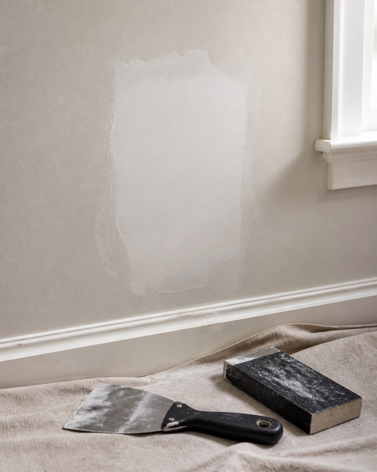 Drywall patch, taping knife, and sanding block beside a Maryland home window
